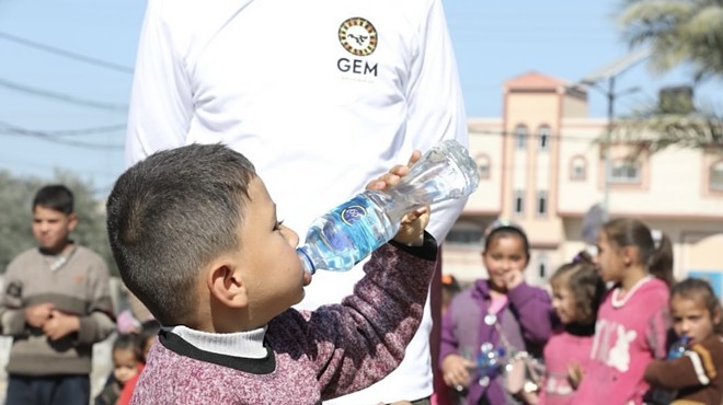 A child in Gaza drinks from a water bottle in front of a person wearing a shirt with a Global Empowerment Mission logo.