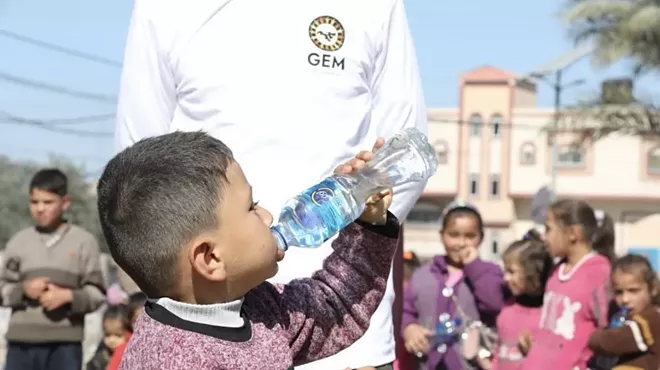 A child in Gaza drinks from a water bottle in front of a person wearing a shirt with a Global Empowerment Mission logo.