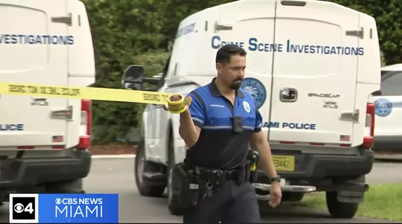 Image: a Miami law enforcement officer pulls crime scene tape across a residential street with police crime scene investigation vehicles in the background
