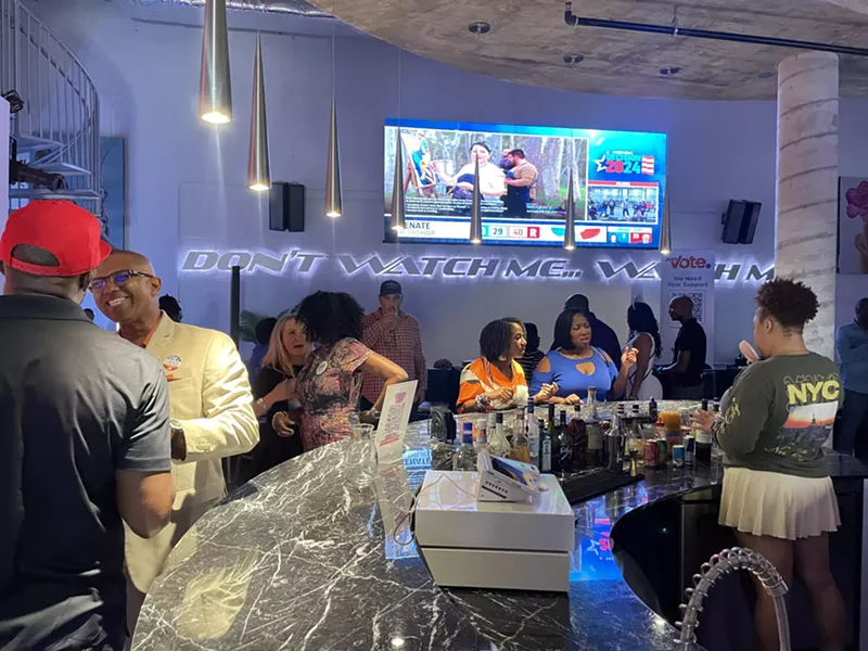 Image: Crowd at Just Vote Period watch party gathered around the bar with a screen displaying election results in the background