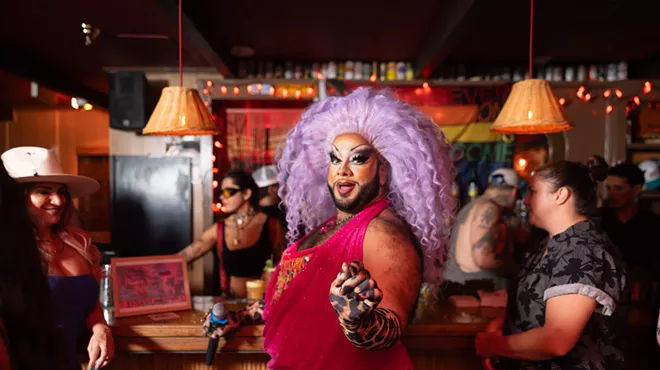 a drag queen in a sparkling pink dress and lavender wig, standing in the middle of a bar