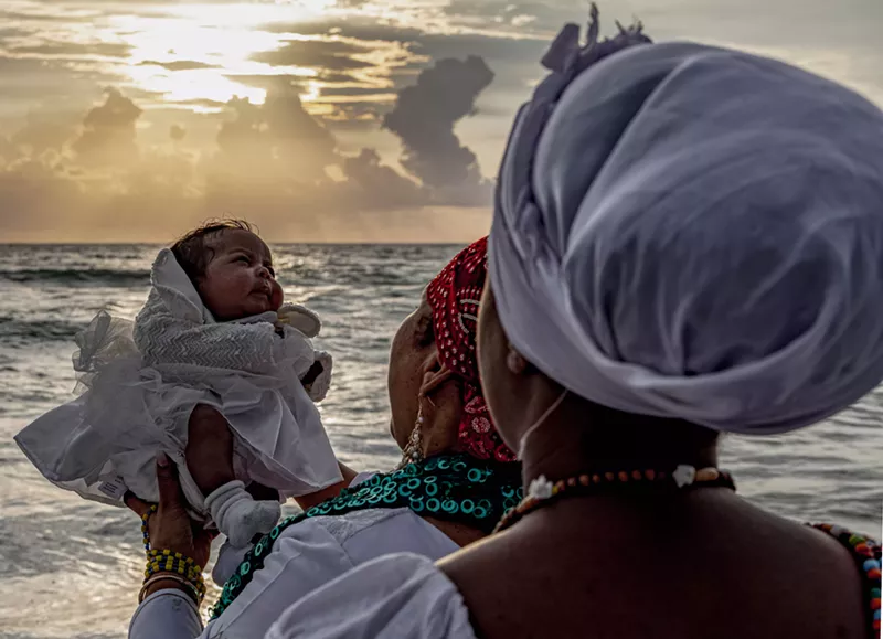 Image: A photo by Woosler Delisfort depicting a spiritual baptism ritual with a child held at the ocean’s edge during sunrise