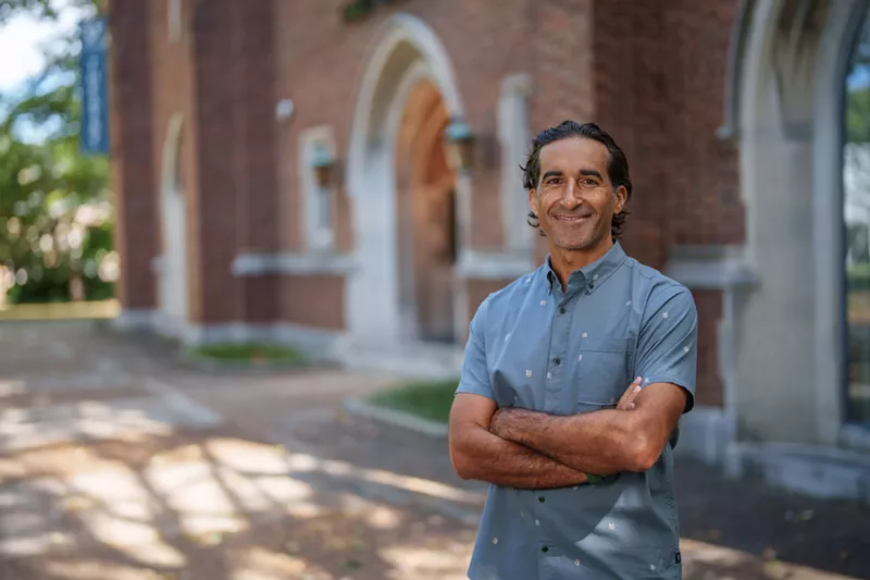 Image: Portrait of a man in a button-down blue shirt standing in front of a brick building