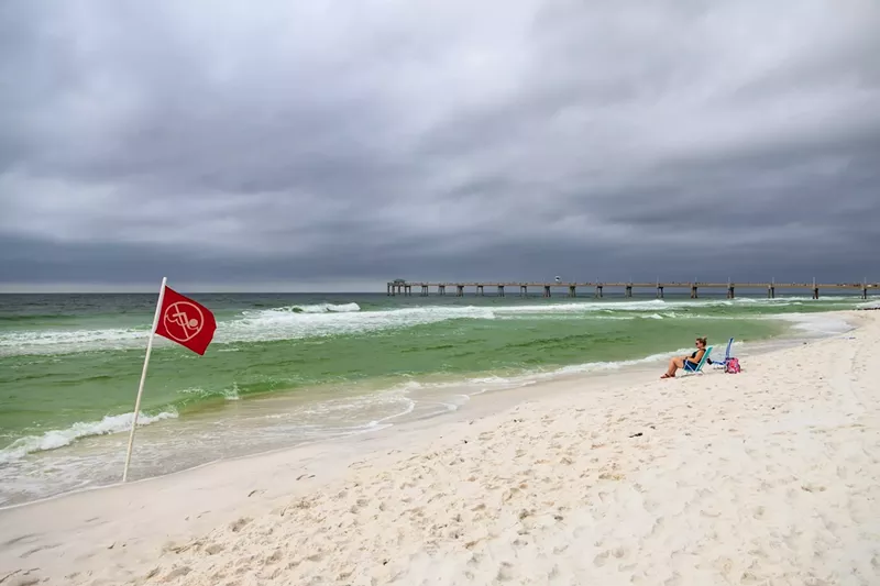 Image: a woman sitting on a beach with stormy weather rolling in; a red flag in the foreground