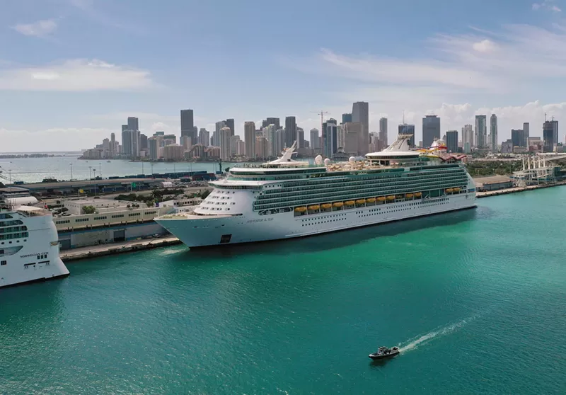 Image: A large cruise ship docked in a port, with the Miami skyline in the background.