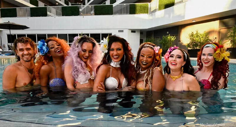 Image: color photo of a row of seven performing mermaids smiling and posing in a swimming pool