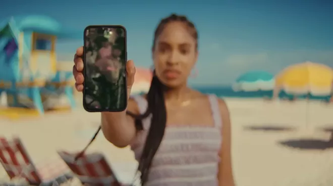A screenshot of a Black female actor with long braids on Miami Beach holding a smartphone up to the viewer