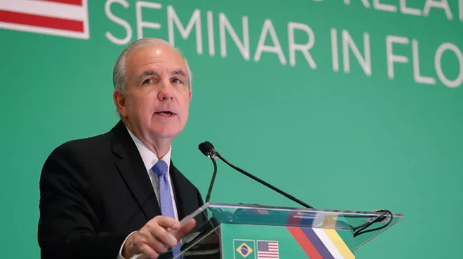 Congressman Carlos Giménez speaks at a podium with a blank green background