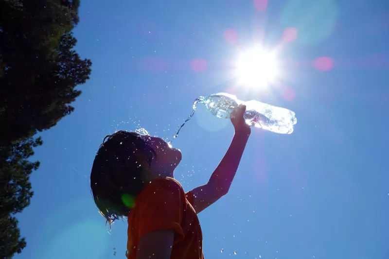 Image: A child drinks from a water bottle with the sun beaming in the background
