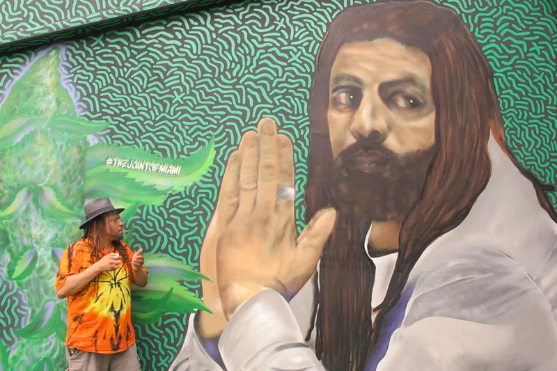 Image: NJWeedman sparks a joint while standing in front of a mural of himself.