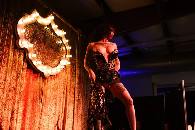 Image: a burlesque dancer lowers a corset to reveal bejeweled tassels on her breasts