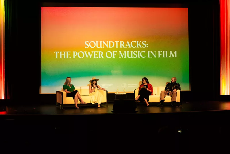 Image: a panel of four people sit on chairs on a stage in front of a projection with the words, "soundtracks: the power of music in film"