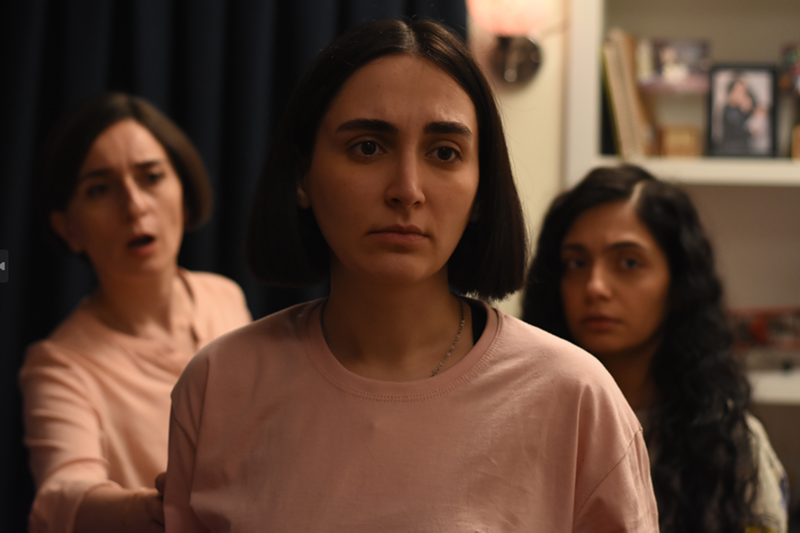 Image: A closeup of a woman in a pink shirt. Behind her, an older woman bears an expression of concern. A young girl in the background on the right is stoic.