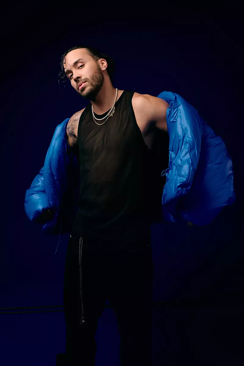 Image: Portrait of a man posing for the camera wearing black pants, a black shirt, and a blue jacket.