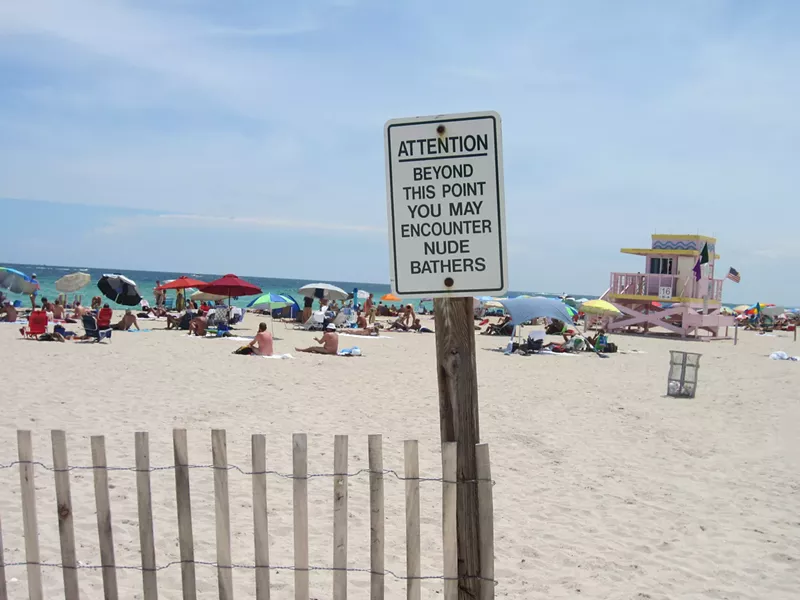 Image: A sign leading to a sandy Miami beach that reads, in all uppercase letters: Attention: Beyond this point you may encounter nude bathers