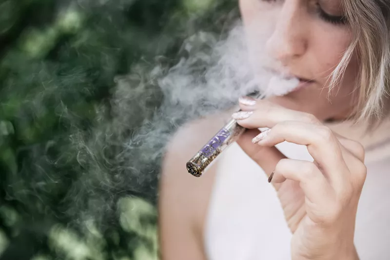 Image: A woman with short hair who is wearing a white tank top puffs on a one hitter pipe packed with what appears to be marijuana while sitting outdoors.