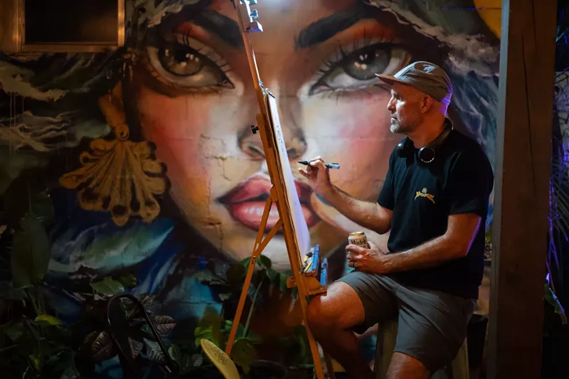 Image: a man paints on a canvas in front of a mural of a woman's face