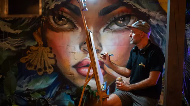 a man paints on a canvas in front of a mural of a woman's face