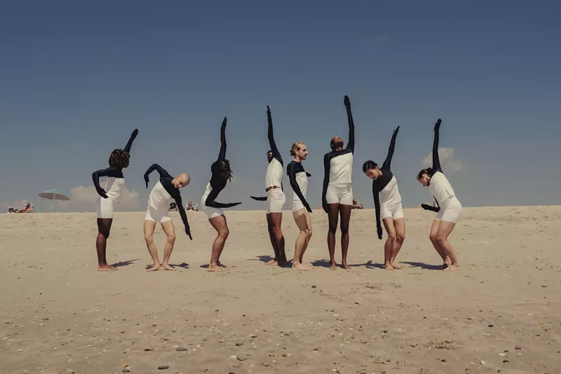 Image: dancers in pose on a beach in white shirts and shorts with arms and hands covered in black cloth