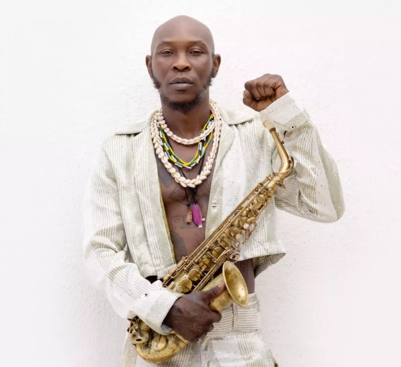 Image: Portrait of a bald, Black man dressed in white with his shirt open, wearing shell necklaces, holding a saxophone, and making a fist