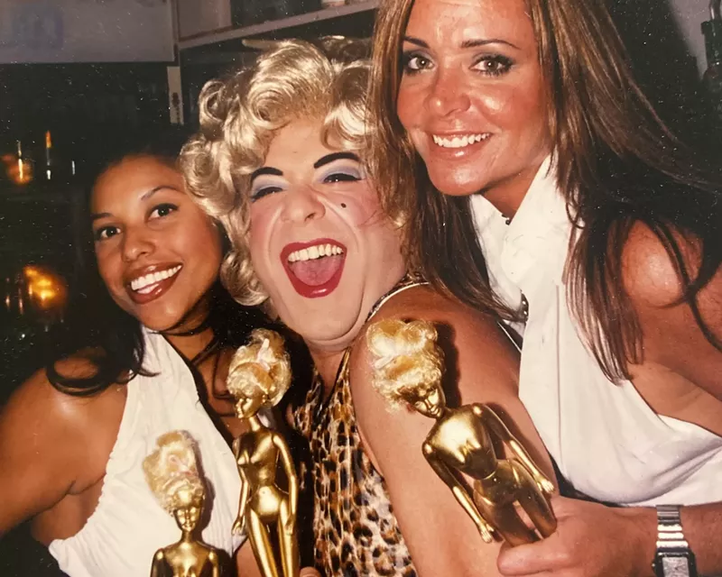 Image: Drag queen Shelley Novak posing alongside two women while holding gold spray-painted Barbies