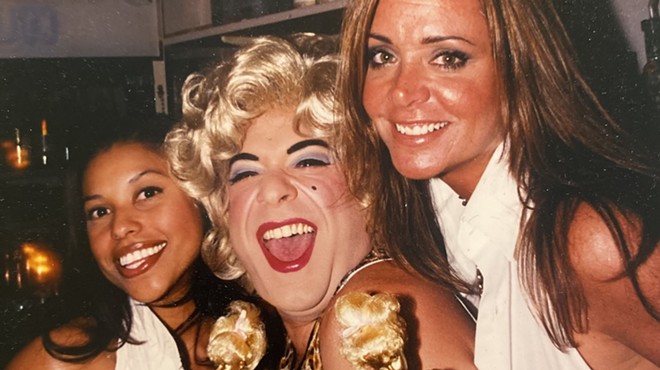 Drag queen Shelley Novak posing alongside two women while holding gold spray-painted Barbies