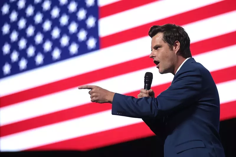 Image: photo of matt gaetz holding a microphone and pointing, presumably to a crowd, against a large american flag backdrop