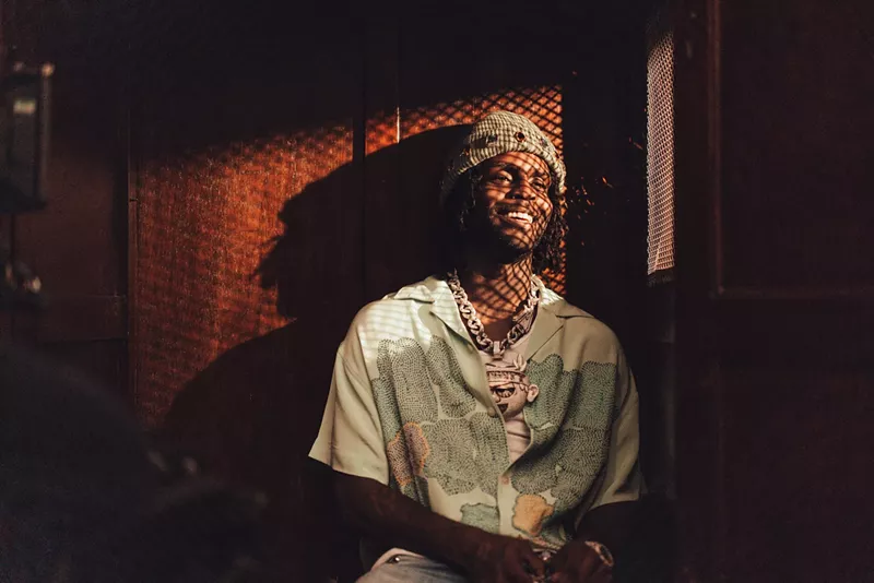 Image: color photo of rapper Chief Keef, smiling, wearing a knit hat and colorful short-sleeved print shirt, and illuminated by sunlight shining through a diamond-patterened mesh screen.