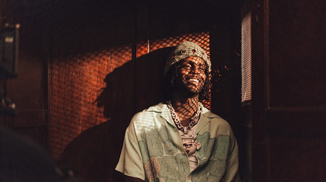 color photo of rapper Chief Keef, smiling, wearing a knit hat and colorful short-sleeved print shirt, and illuminated by sunlight shining through a diamond-patterened mesh screen.