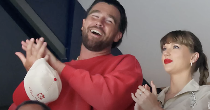 Image: A man in a red sweater and a woman in a khaki top and red lipstick clap and smile