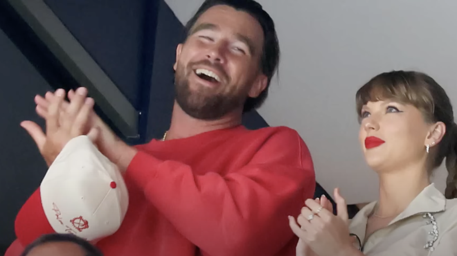 A man in a red sweater and a woman in a khaki top and red lipstick clap and smile