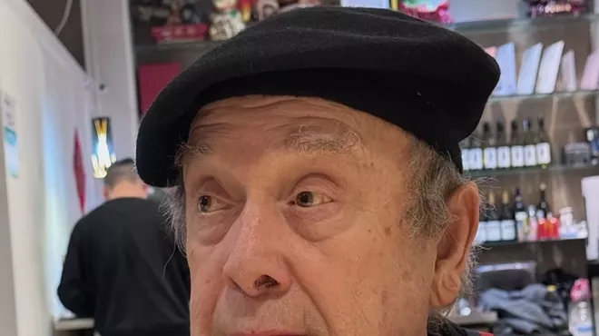 Photo of a 79-year-old man wearing a black beret and a black short-sleeve shirt with red-and-black suspenders, with his hands clasped under his chin.