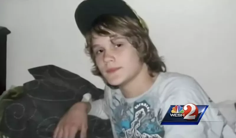 Image: color snapshot of a young boy with shaggy shoulder-length brown hair, wearing a t-shirt and a black ball cap perched high on his head, looking into the camera