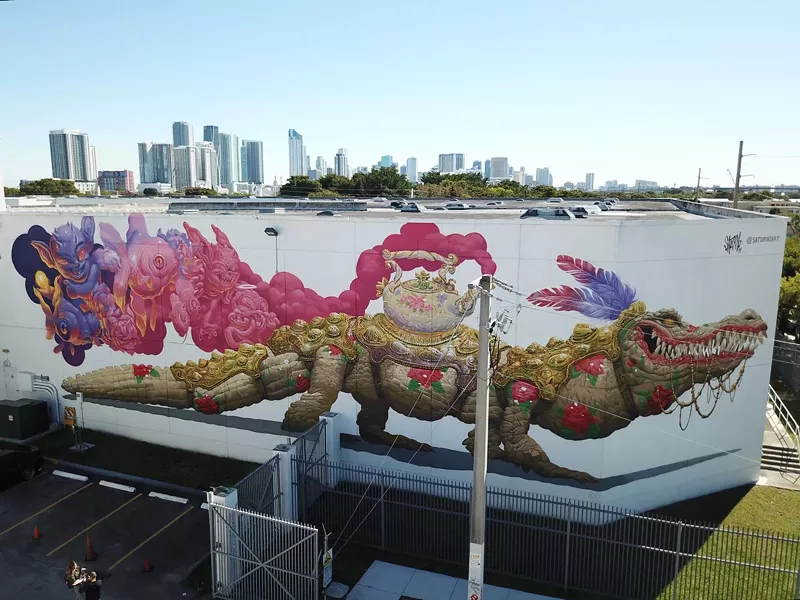 Image: a white building displays a mural with an alligator wearing regal decor, a tea kettle, and feathers