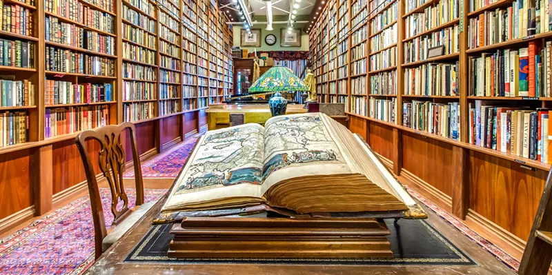 Image: An open book on a table shows a map. Behind it are an antique lamp and bookshelves with hundreds of books