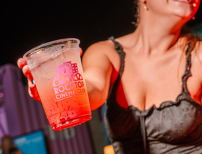 Image: a woman holds up a plastic cup with a cocktail reading, "Rooftop Cinema Club"