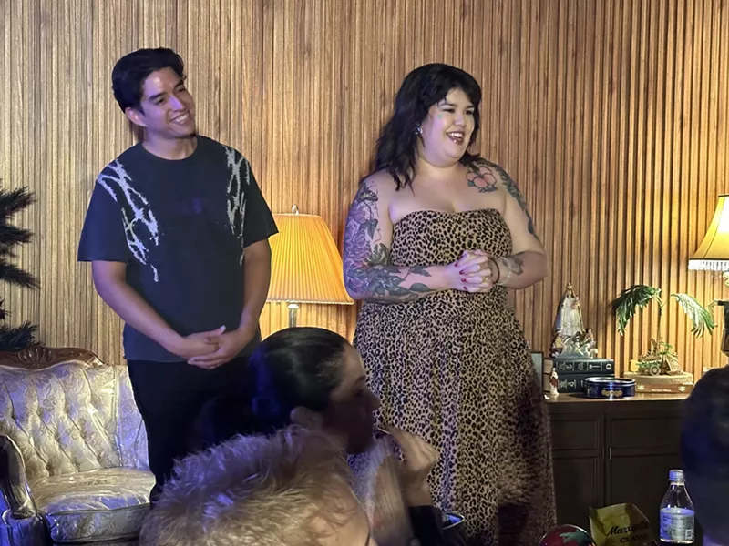 Image: a performer speaks to an audience in a living room as another performer stands next to her