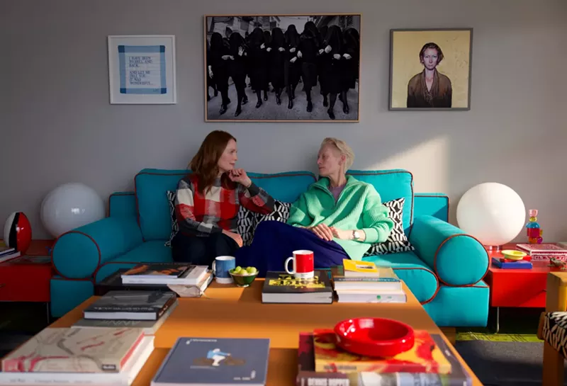 Image: Still of Tilda Swinton and Julianne Moore sitting on a teal couch. Framed photos hang above the couch