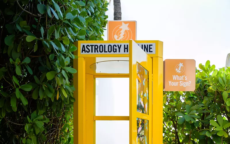 Image: yellow old-school telephone booth. a sign on the booth reads "astrology hotline." another sign protruding from its side reads "what's your sign"