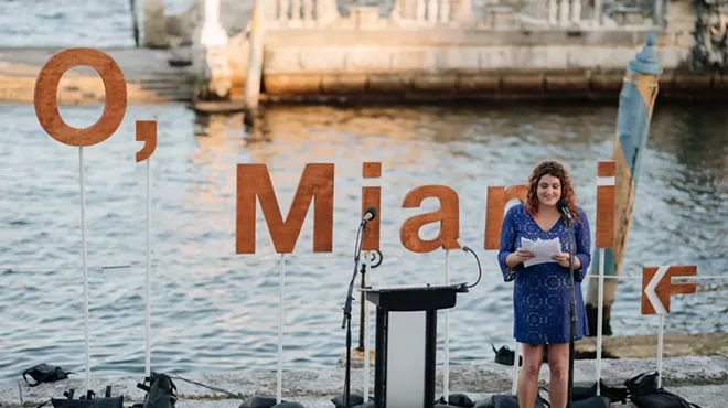 a person reads a poem in front of the water and a backdrop that reads, "O, Miami"