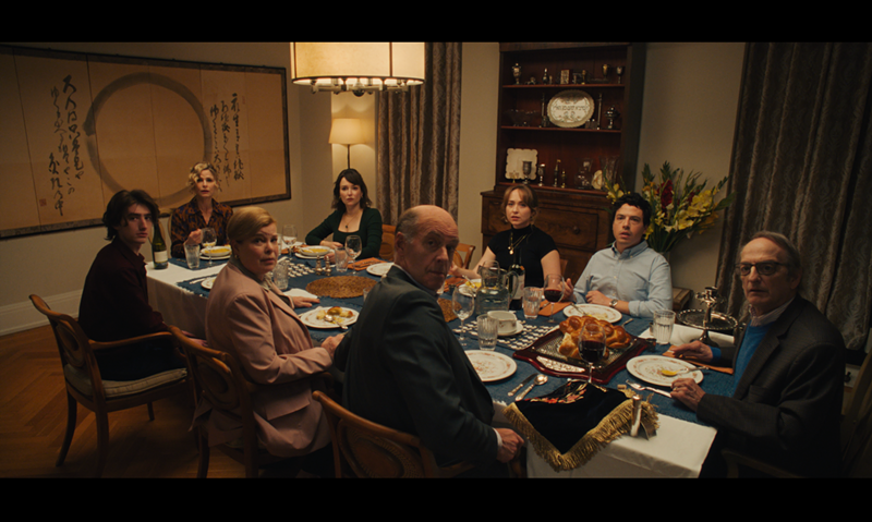 Image: a film still of eight family members gathered around the dinner table glaring at the camera in unison