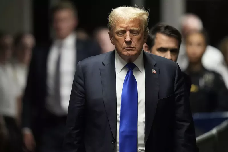 Image: photo of Donald Trump wearing blue suit, white shirt, and blue(!) tie, walking, looking somber, with his eyes nearly closed