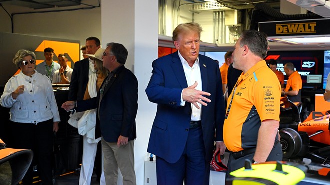 Donald Trump stands in front of a race car, talking to a McLaren executive while gesturing with his right hand