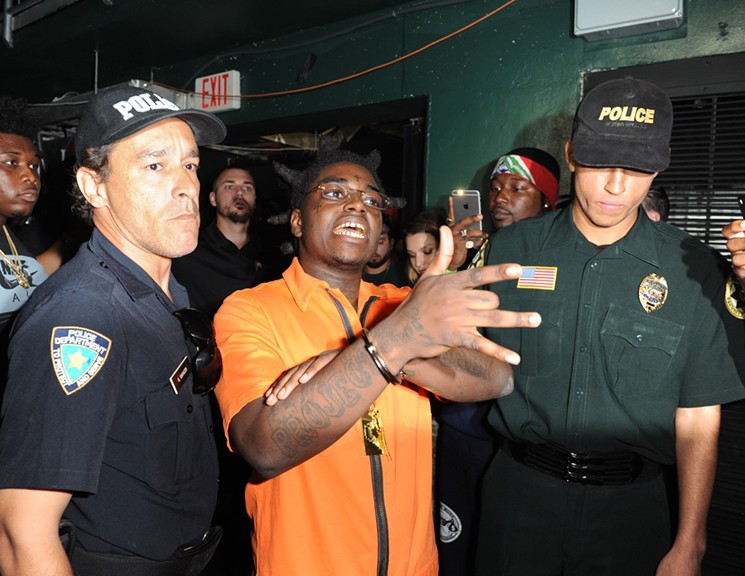 Kodak Black in a posed color shot, dressed in an orange prison jumpsuit, standing between two white men in police gear