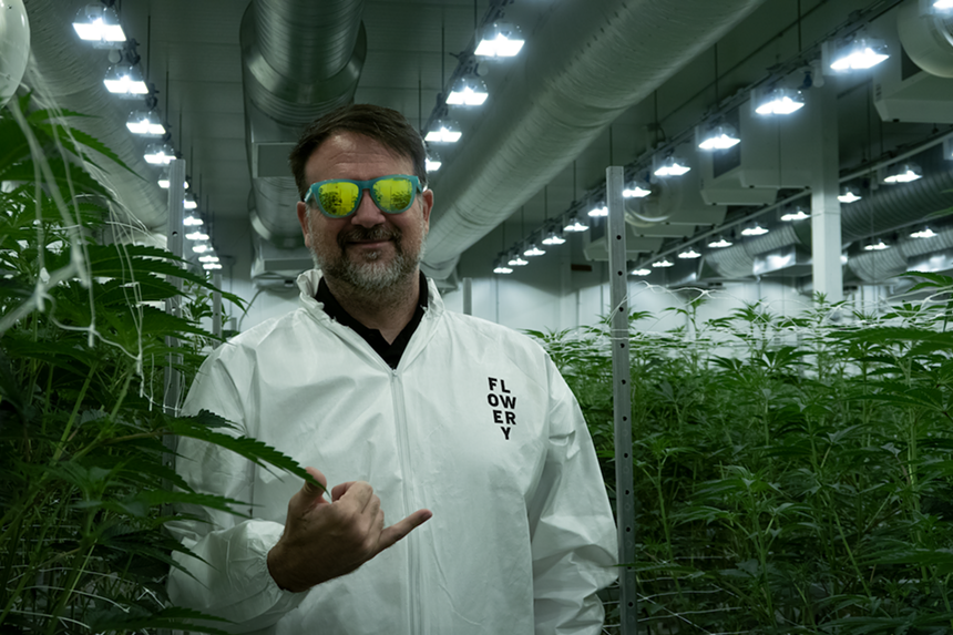 a bearded, white-coated man wearing mirrored sunglasses stands in a greenhouse filled with marijuana plants