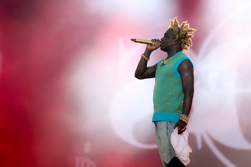 color photo of Kodak Black wearing a teal sleeveless shirt, holding a microphone, and singing, against a pink spotlit background
