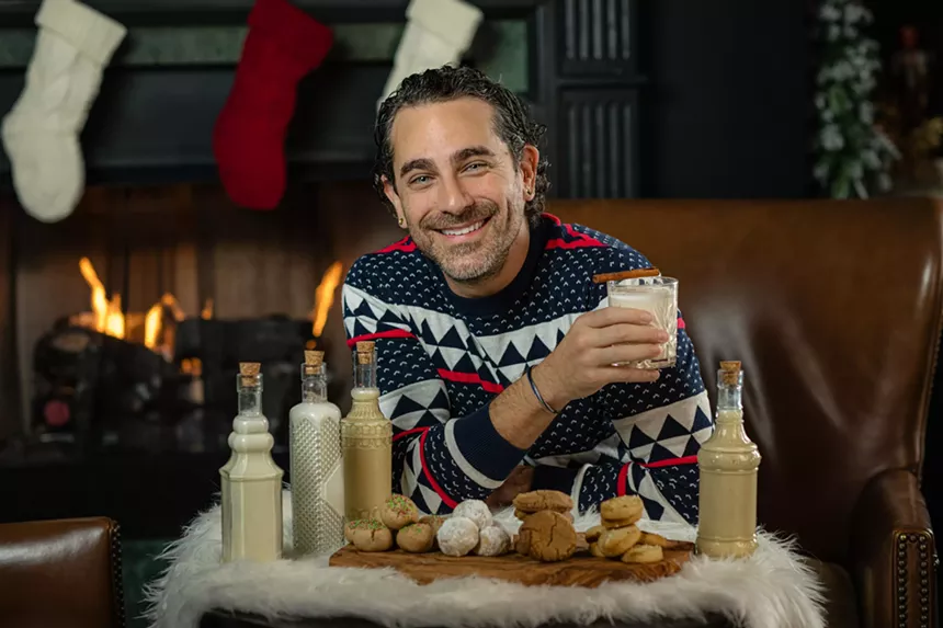 Gio Gutierrez of ChatChowTv poses with his coquito creations and their cookie pairings that will be offered at the Ritz-Carlton Coconut Grove this December. He is wearing a Christmas sweater and is holding a holiday drink with a cinnamon stick laid on top.