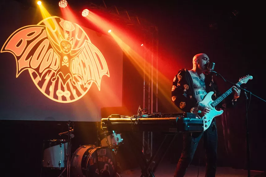 Miami musician Niko Javan singing into a microphone and strumming his guitar, performing as Blood Orchid, as a keyboard sits nearby