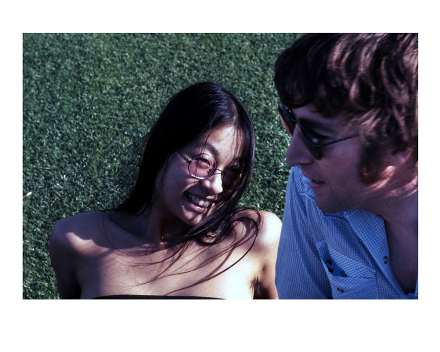 Color photo of a sun-dappled May Pang lying on the grass next to a bespectacled John Lennon