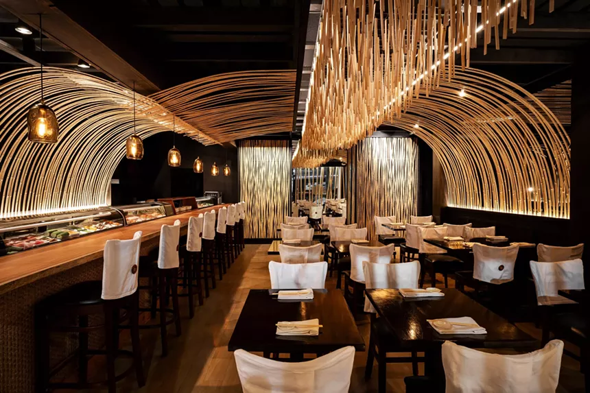 bamboo-accented restaurant interior with tables in the center and a sushi bar on the left that's illuminated by hanging lamps